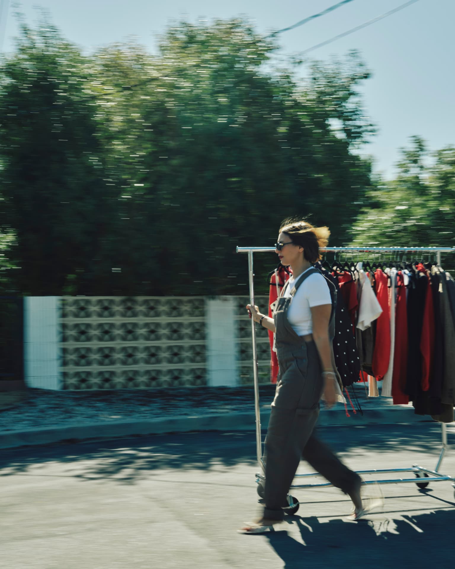 A woman in sunglasses pushing a rolling rack of clothing down a sunlit street