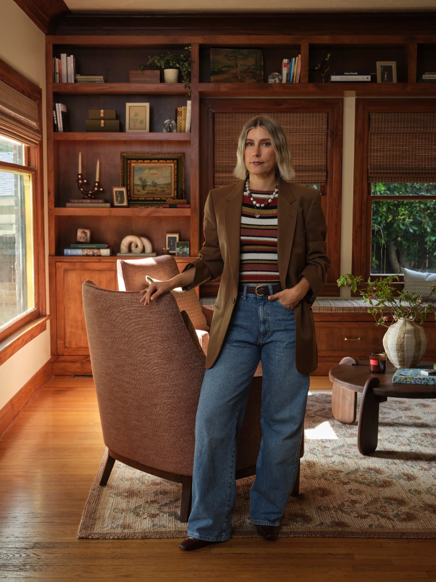 A woman in a camel blazer and striped knit standing in a wood-lined library, one hand resting on a high-backed armchair